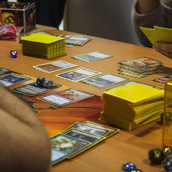 Billedet viser et bord, hvor folk spiller magic the gathering på. Der ligger kort på bordet, man kan se to personer og der er et deck med gule sleeves til hver person.