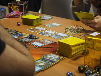 Billedet viser et bord, hvor folk spiller magic the gathering på. Der ligger kort på bordet, man kan se to personer og der er et deck med gule sleeves til hver person.