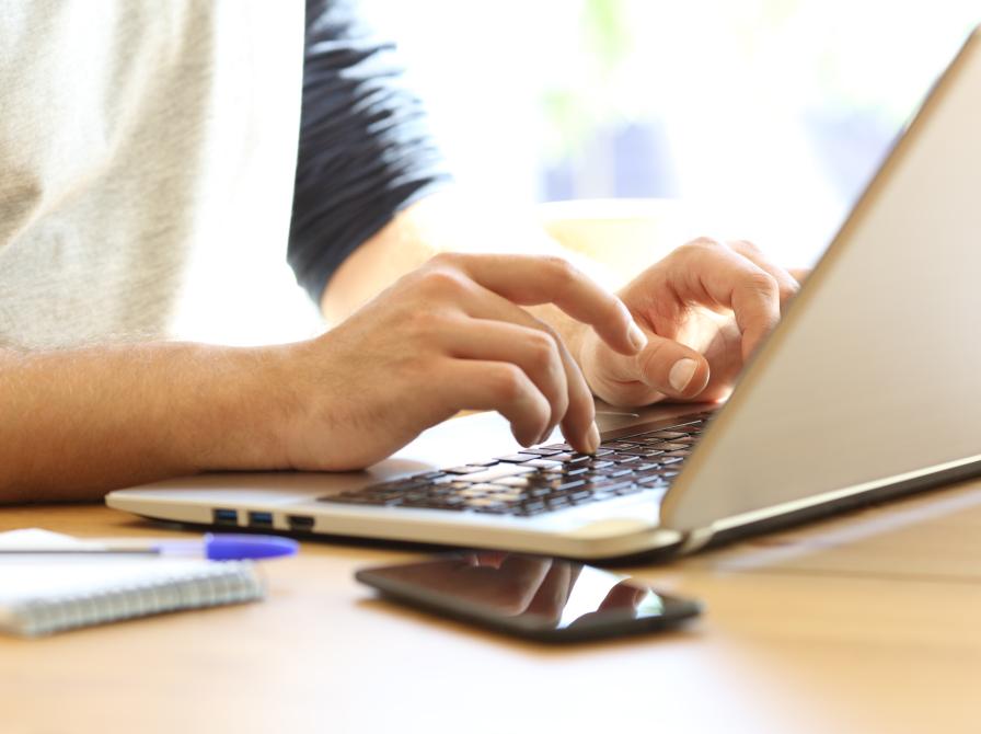 Bærbar computer, notesblok og telefon på et bord og hænder, der skriver på pc'en